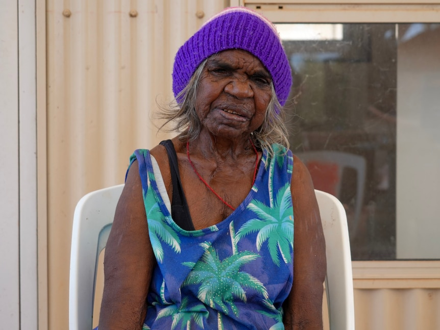 An Aboriginal female elder stares front on wearing a purple beanie.
