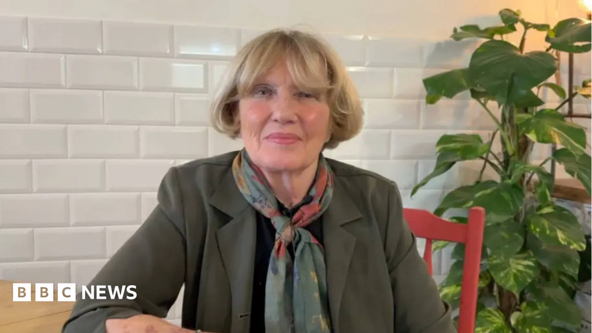 Denise Mackay with blonde hair, wearing an olive blazer and a pashmina. She is sitting at a wooden table on a red chair looking at the camera.