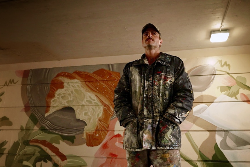 A man standing in front of a mural painted on a wall in a tunnel looking straight ahead.