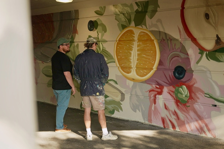 Two men stand in a tunnel looking at a brightly coloured mural painted on a wall.