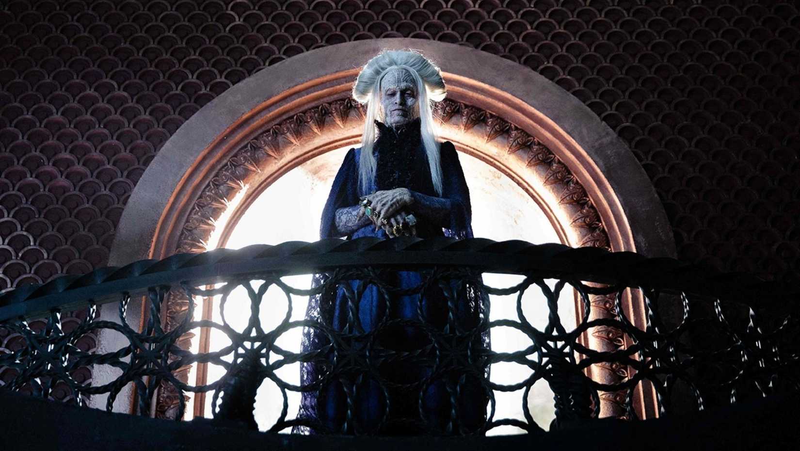 In a scene from Luc Besson's Dracula, Caleb Landry Jones, playing the title monster, stands on an ornate balcony in front of a window, wearing his white hair "up" in a way that looks like Gary Oldman's version of the character.