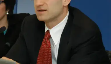 A man in a dark suit, white shirt, and a red patterned tie, wearing glasses, sits at a table. He is looking off to the left with an attentive expression. A silver laptop is in the foreground on the left, and a partial view of a plastic water bottle is on the right. Papers and a pen are visible on the table. A blue wall forms the background, and another person is partially visible on the far left.
