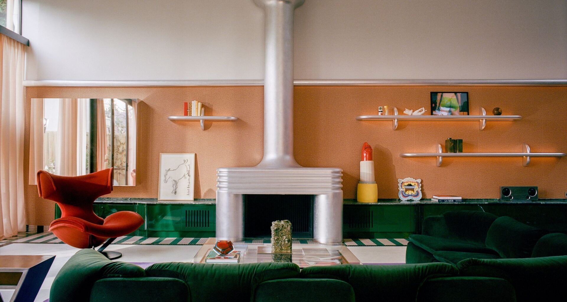 Image of a large living room with orange wallpaper and white paint in a color block, a green, glossy built-in, low cabinet, a green sofa, a chrome fireplace, and a red chair.