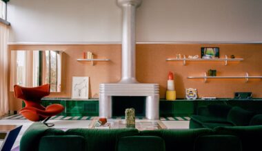 Image of a large living room with orange wallpaper and white paint in a color block, a green, glossy built-in, low cabinet, a green sofa, a chrome fireplace, and a red chair.