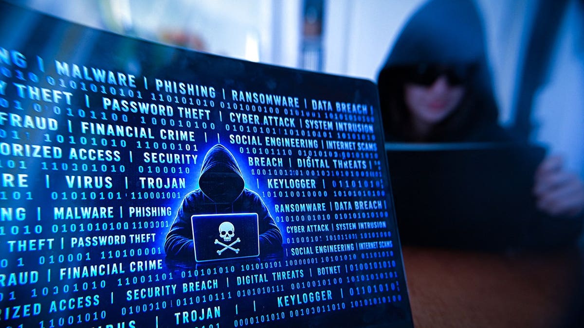 Hooded person uses a digital tablet beside a laptop displaying a pirate symbol in a dark setting.
