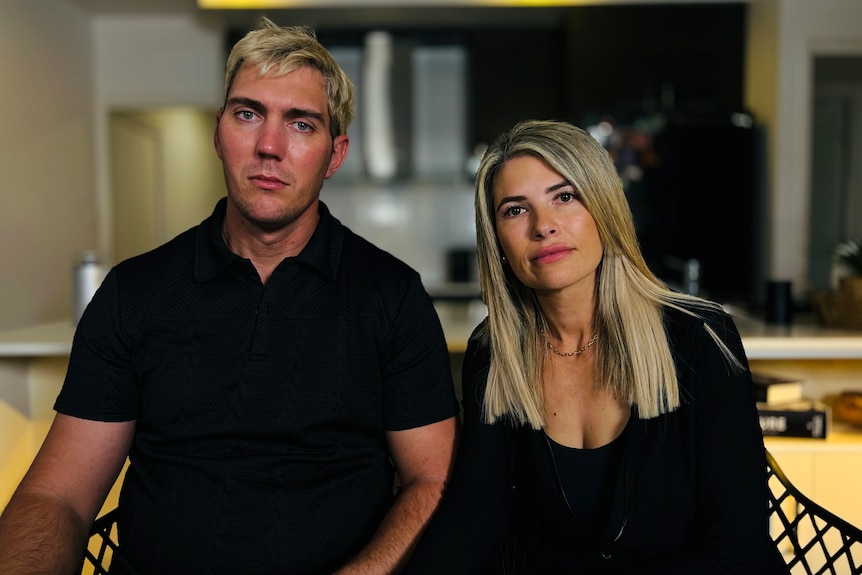 A man and a woman sit in an apartment. They are looking at the camera with serious expressions.