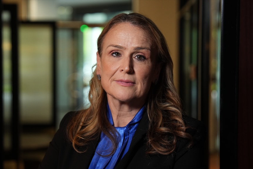 Misha Schubert, a woman with light brown hair, wears a black jacket over a blue shiny top with a serious facial expression.