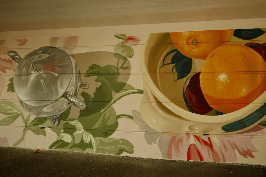 A mural on a wall in a tunnel showing a silver pot and two oranges in a bowl.
