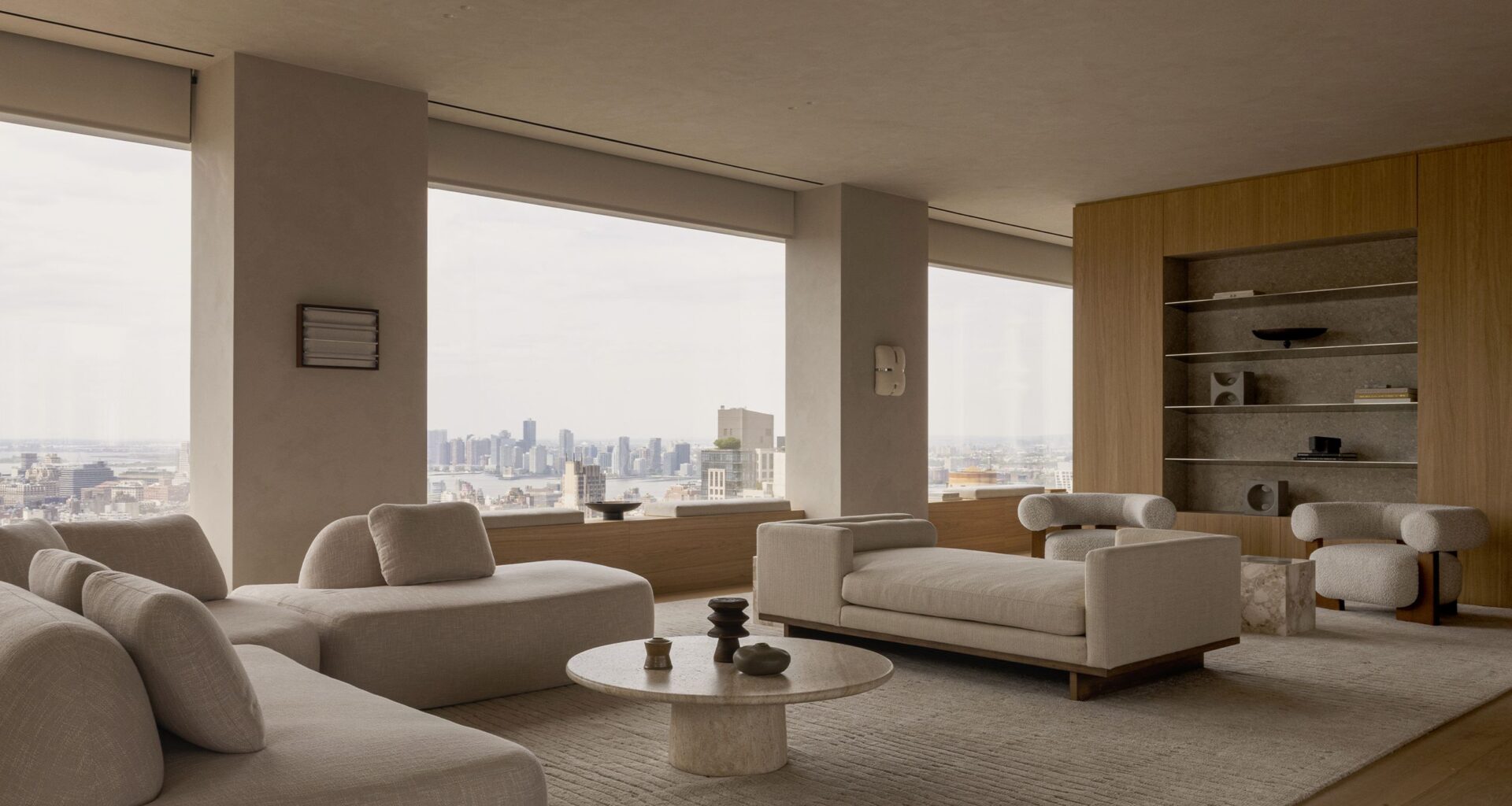 Neutral-toned living room with floor-to-ceiling windows overlooking the Manhattan skyline, featuring sculptural seating, natural oak cabinetry and a stone coffee table.