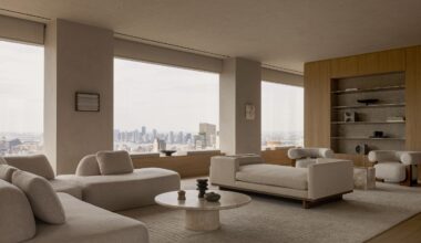 Neutral-toned living room with floor-to-ceiling windows overlooking the Manhattan skyline, featuring sculptural seating, natural oak cabinetry and a stone coffee table.