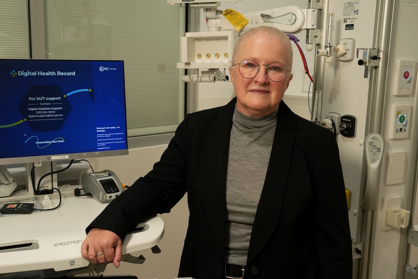 A woman with grey hair in a ponytail stands in a hospital room looking serious.