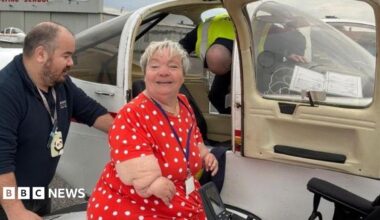 Theresa has short grey hair and wears a red polka dot dress. She is pictured standing next to a small plane, smiling. A man appears to be helping her climb into the plane