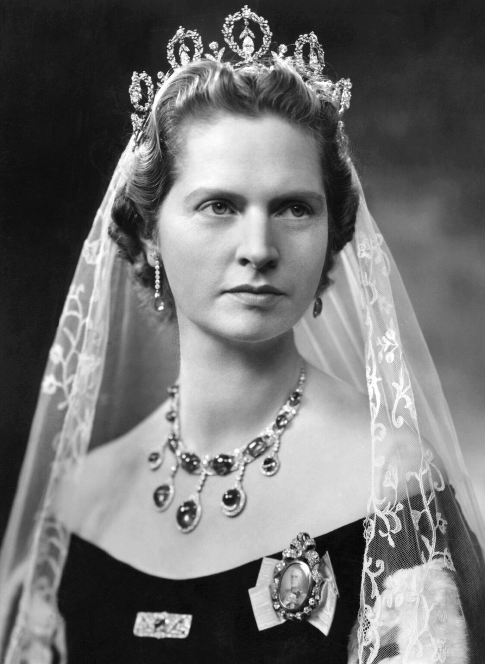 Black and white portrait of a woman wearing a tiara, lace veil, and ornate jewelry, with a serious expression.
