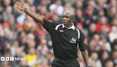 A man wearing a black football shirt stands in front of an out of focus crowd. His left arm is raised. He is wearing an ear piece and microphone on the right side of his face. He has a serious expression.