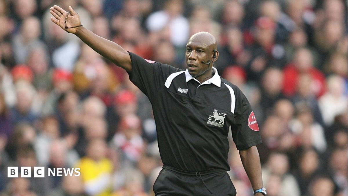 A man wearing a black football shirt stands in front of an out of focus crowd. His left arm is raised. He is wearing an ear piece and microphone on the right side of his face. He has a serious expression.