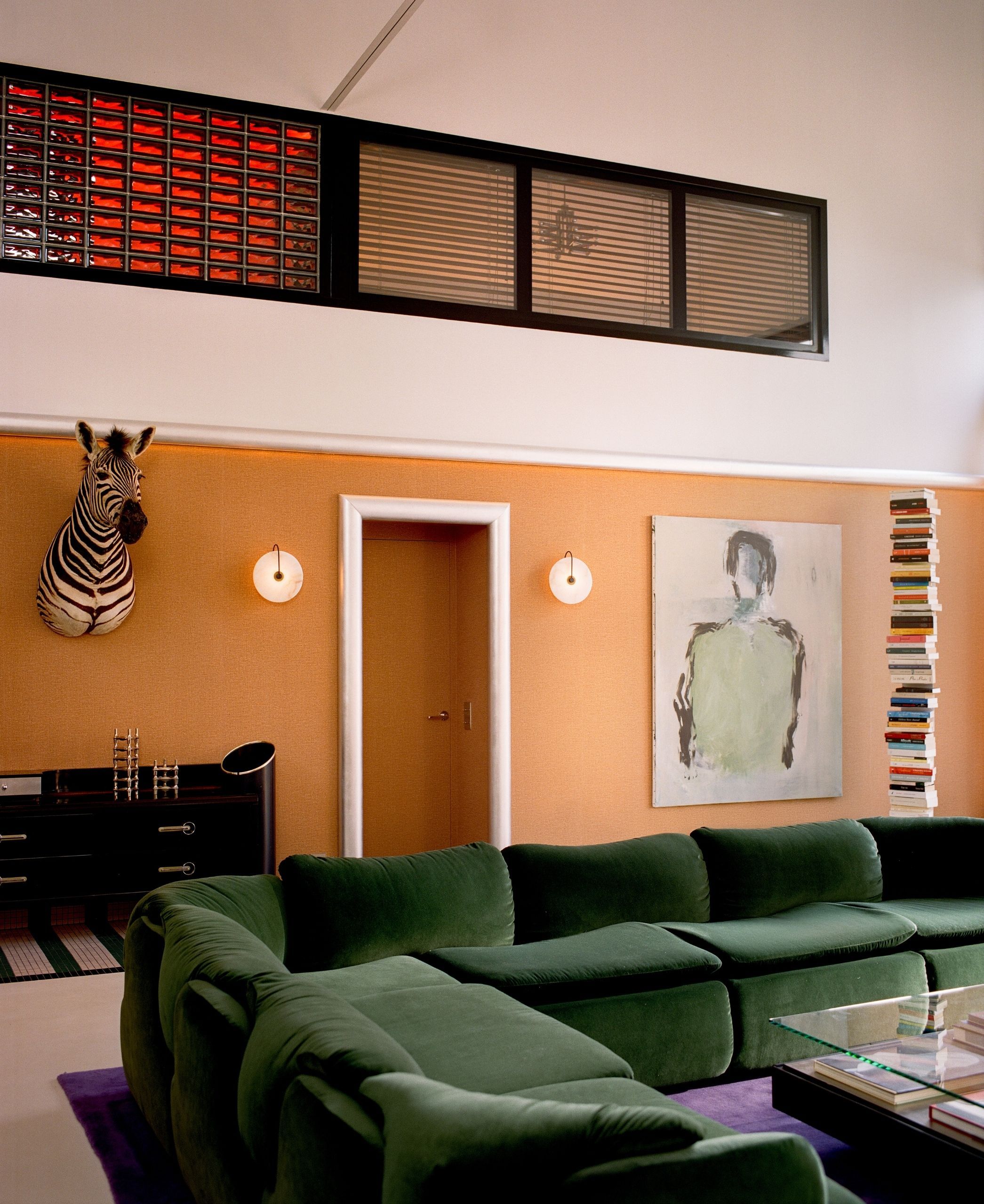 Image of a living room with orange wallpaper and white paint on the walls in a color block. There is a large, dark green sectional sofa and a purple rug. There is a zebra head mounted on the wall.