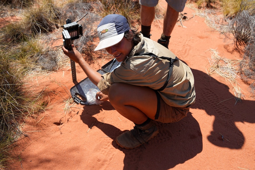 Female ranger squats down in red dirt as she changed batteries from a camera