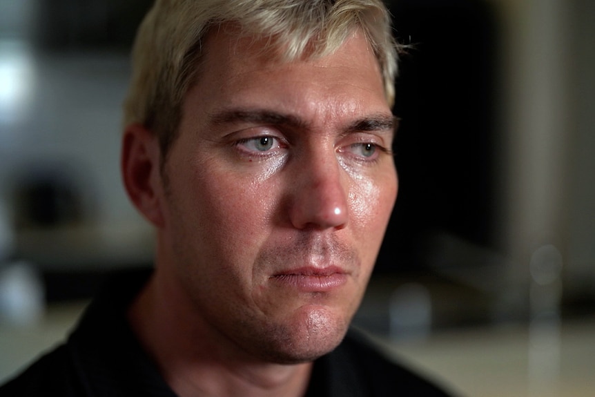 A close-up of a man sitting indoors, looking ahead. His expression is sad.