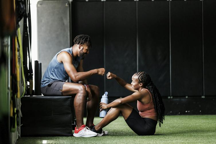 Two people sitting inside a gym, fit bumping