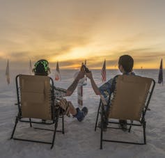 Two people sit in beach chairs on snow clinking bottles with the sun coming over horizon.