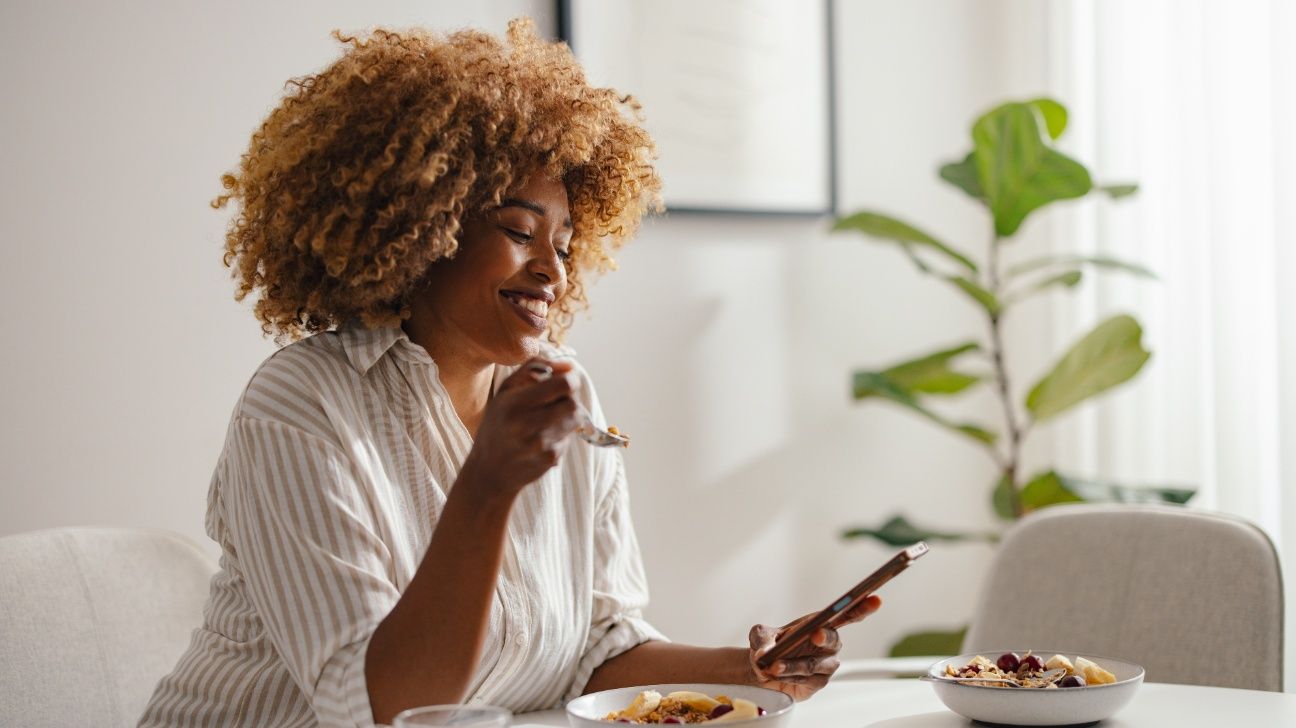 Female sitting at a table, eating a healthy breakfast and scrolling on her phone
