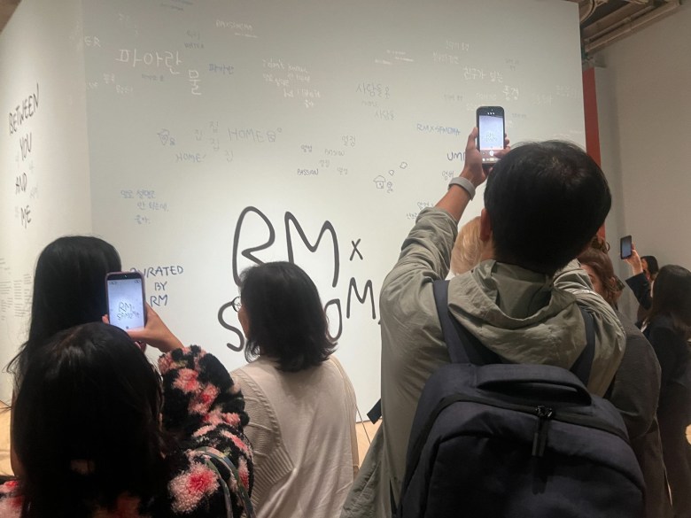 A group of people stand in front of a white wall with “RM x S.R.ROOM” written on it, taking photos with their phones. Handwritten words in various languages are scattered across the wall.