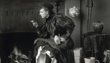 Woman in dark clothing sits by fireplace in vintage interior setting