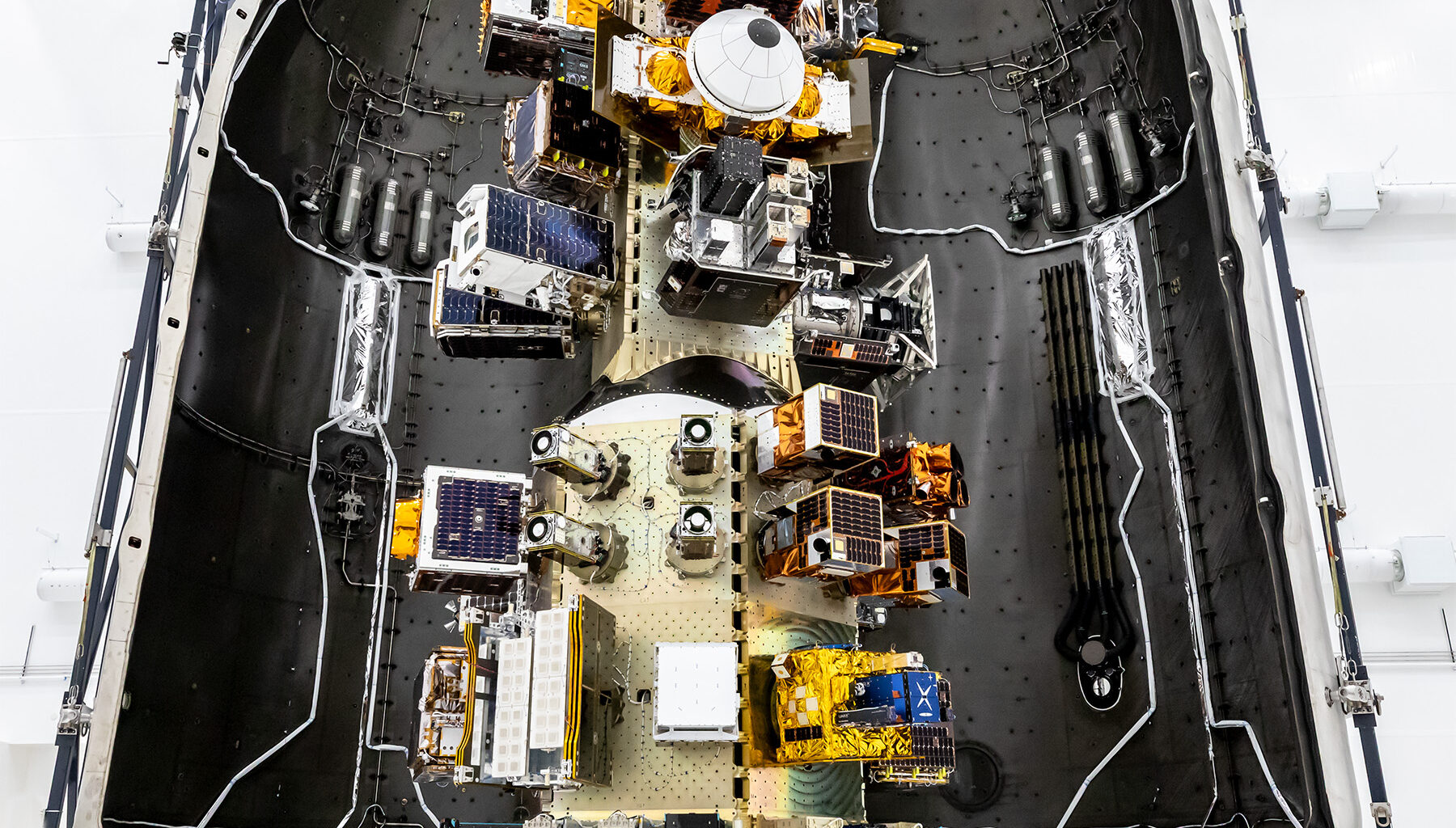 View of a SpaceX payload fairing interior showing mounted satellites and hardware arranged along the center structure, surrounded by the curved black fairing walls.