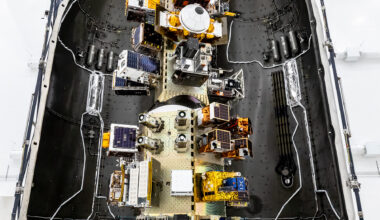 View of a SpaceX payload fairing interior showing mounted satellites and hardware arranged along the center structure, surrounded by the curved black fairing walls.