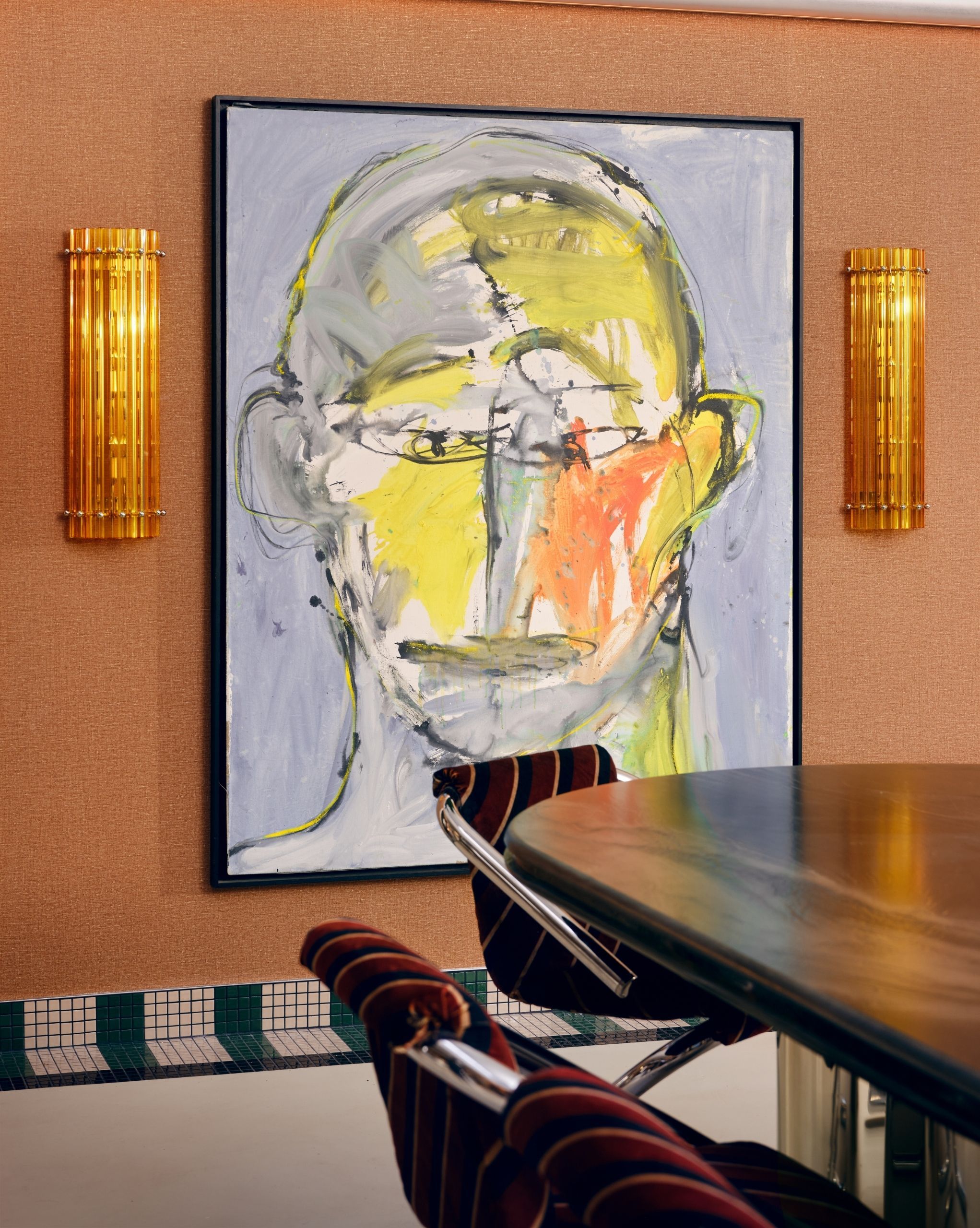 The wall of a dining room covered in orange wallpaper with a giant, abstract painting of a face hung on it. The edge of the table and three chairs are in view.