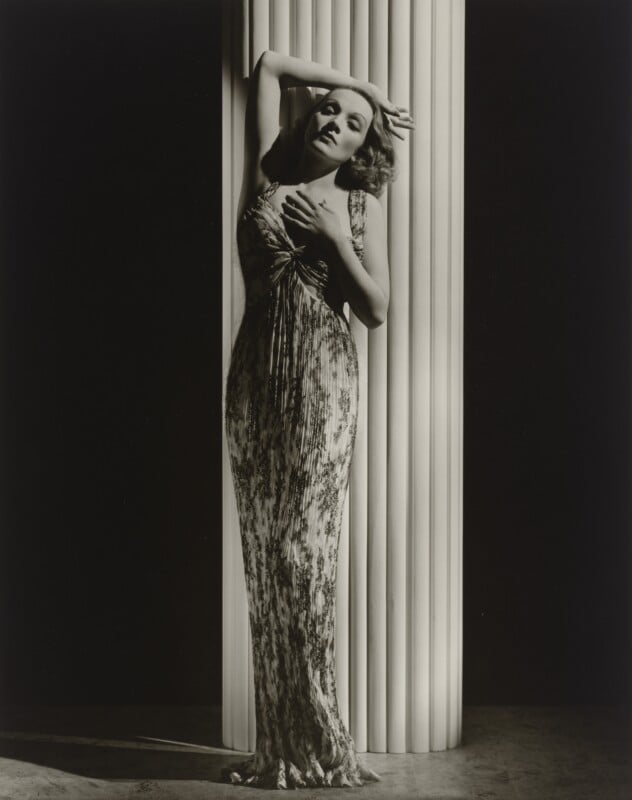 A woman with wavy hair poses against a large, fluted white column, wearing a long, patterned, sleeveless gown.