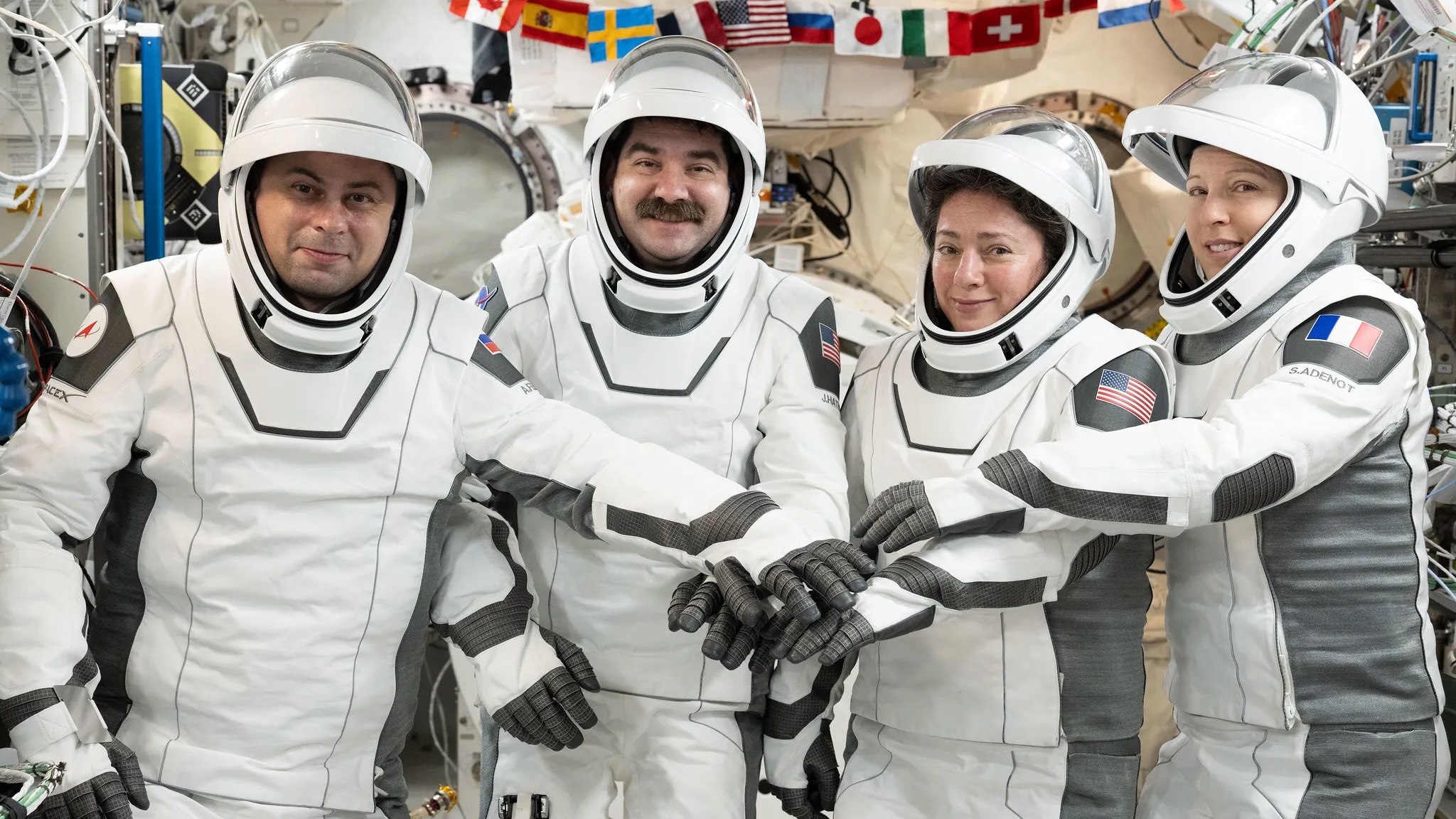 two male and two female astronauts, all wearing white and gray pressure suits, pose inside a space station module