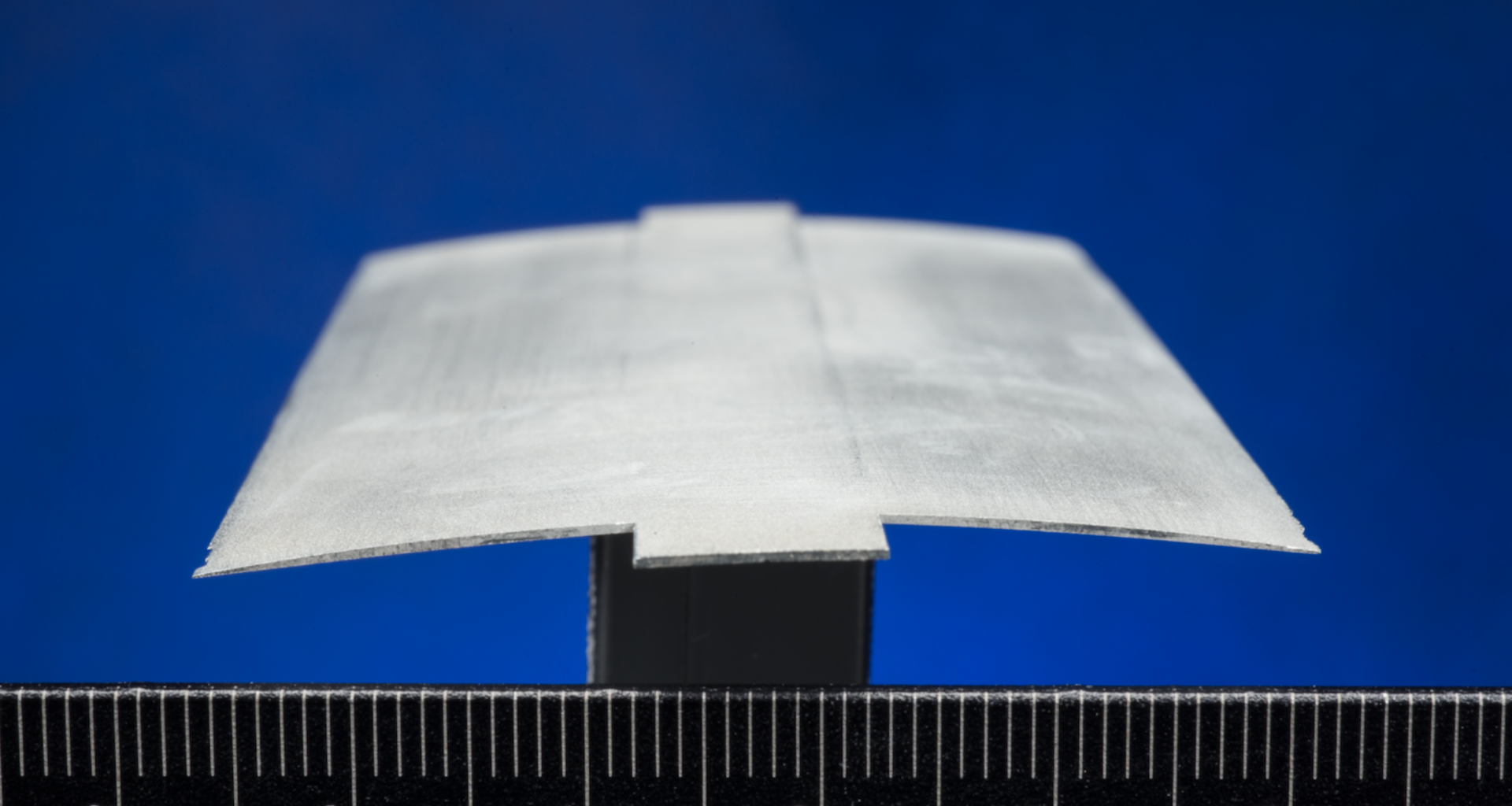 A close-up view of a cross section of a rotor blade, also called an airfoil. The thin, gray material is slightly arched and sits atop a black stand against a ruler.