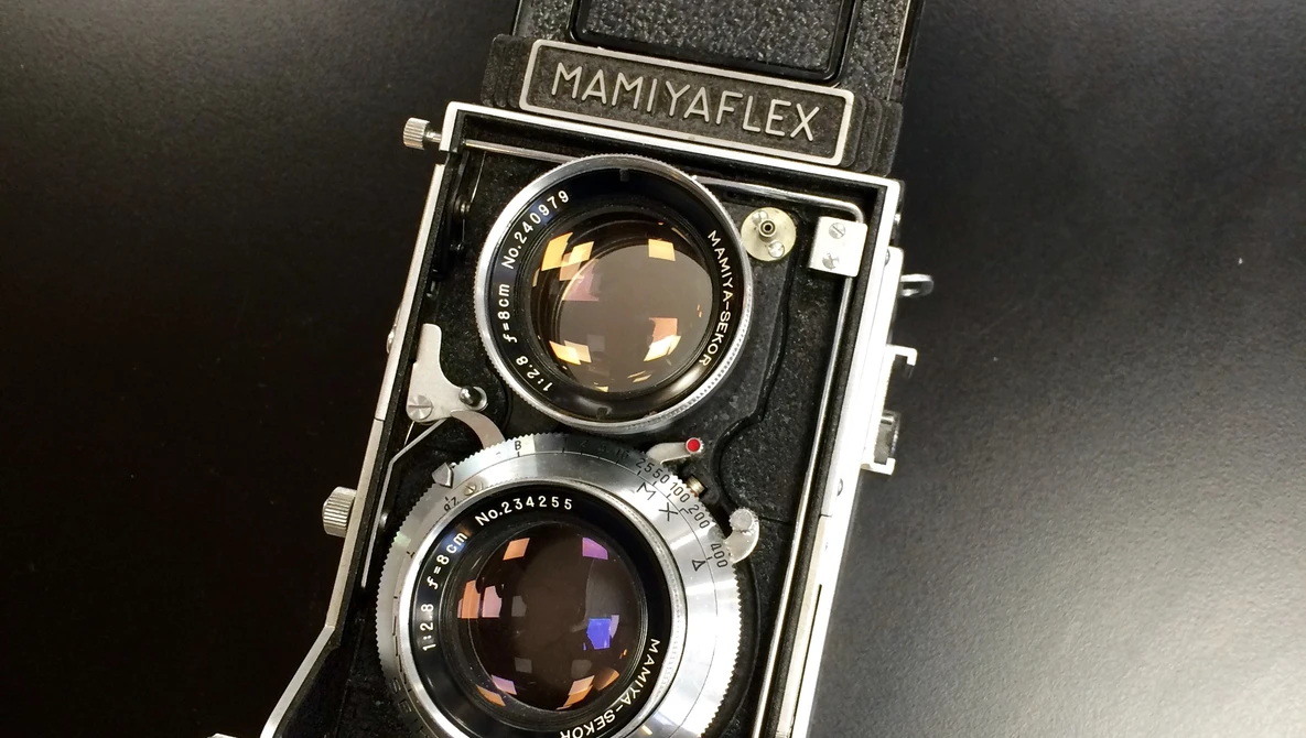 Vintage Mamiyaflex twin-lens reflex camera with a black body, chrome lens barrels, and waist-level viewfinder hood, resting on a dark surface.