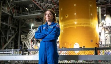 a woman in a blue flight suit stands in front of a large orange rocket inside a large building