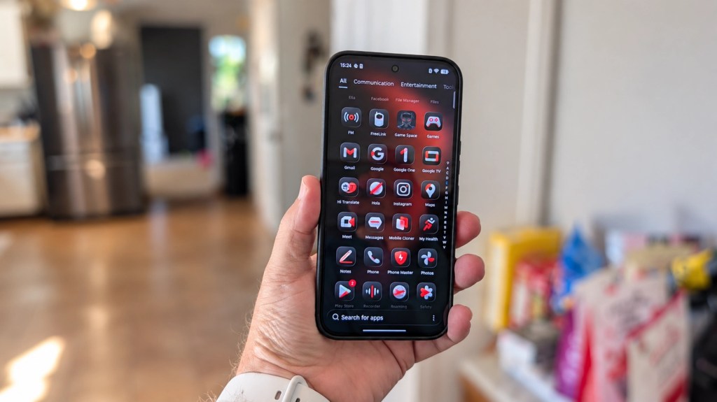A hand holding a smartphone displaying a dark-themed app interface with various applications, set in a kitchen with blurred background elements.