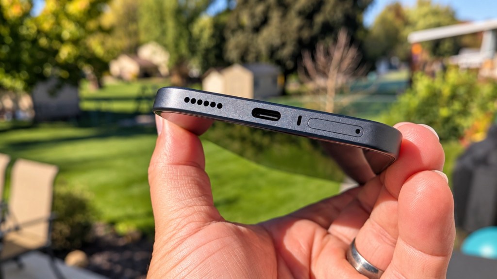 Side view of a smartphone being held, showing the charging port, speaker grill, and SIM card tray, with a blurred green outdoor background.