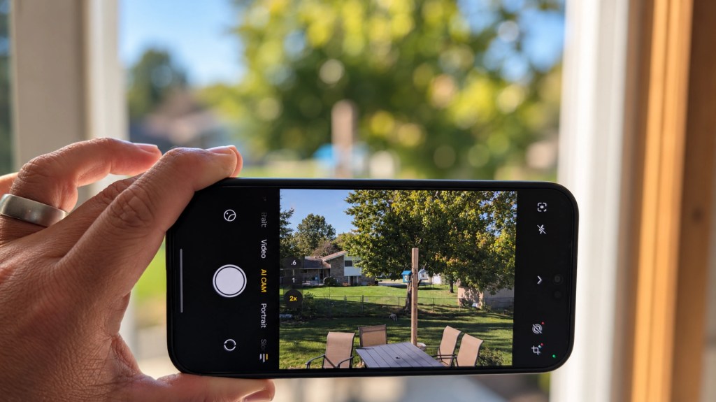 A hand holding a smartphone displaying a photo of an outdoor scene with trees, a table, and chairs.