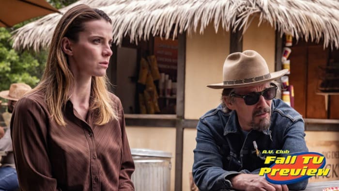 In a still from The Lowdown season 2, Betty Gilpin as Ginger wears a brown shirt and sits next to Ethan Hawke as Lee Raybon wearing a cowboy hat, sunglasses, and a denim shirt. They sit in front of a tropical-themed food truck with faux-thatched awning