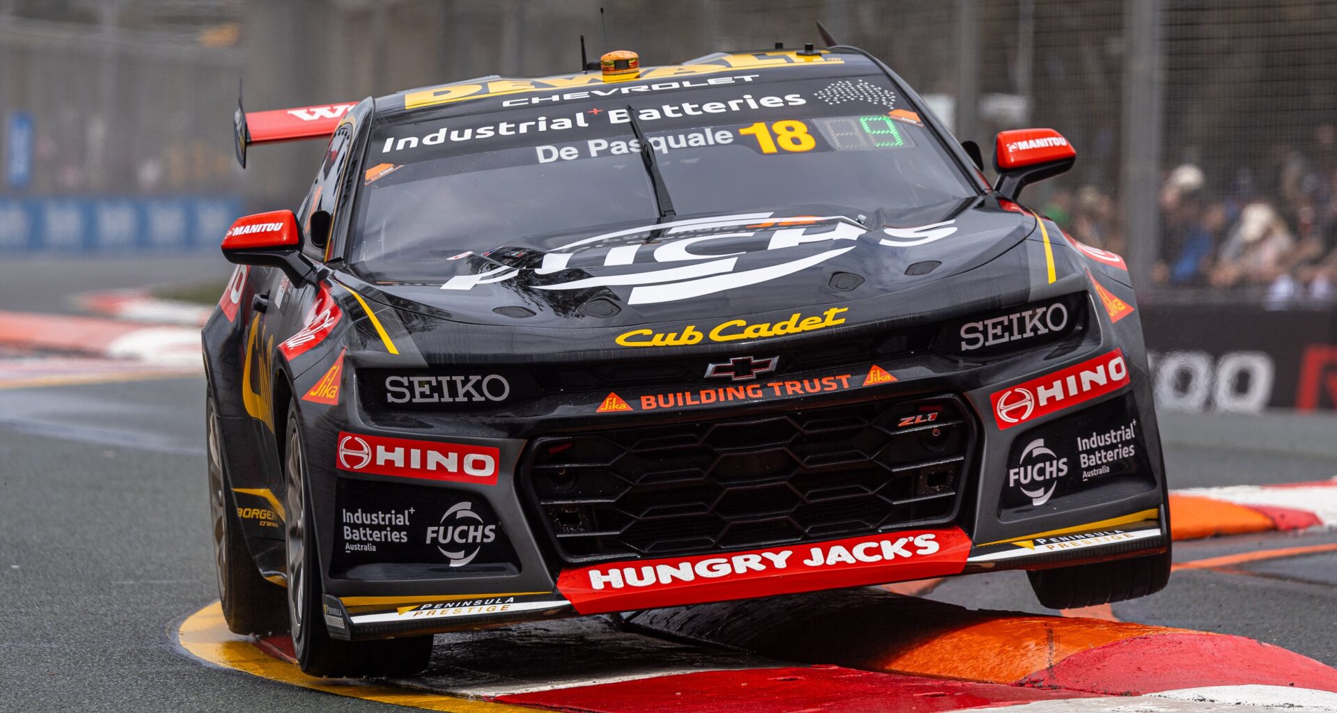 Black and gold Supercar number 18 bounces over a curb at a street circuit.