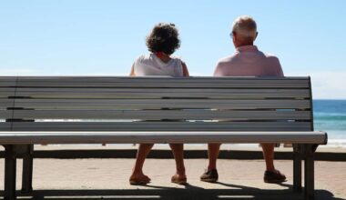 An older couple sit on a bench facing the ocean as seen from behind.