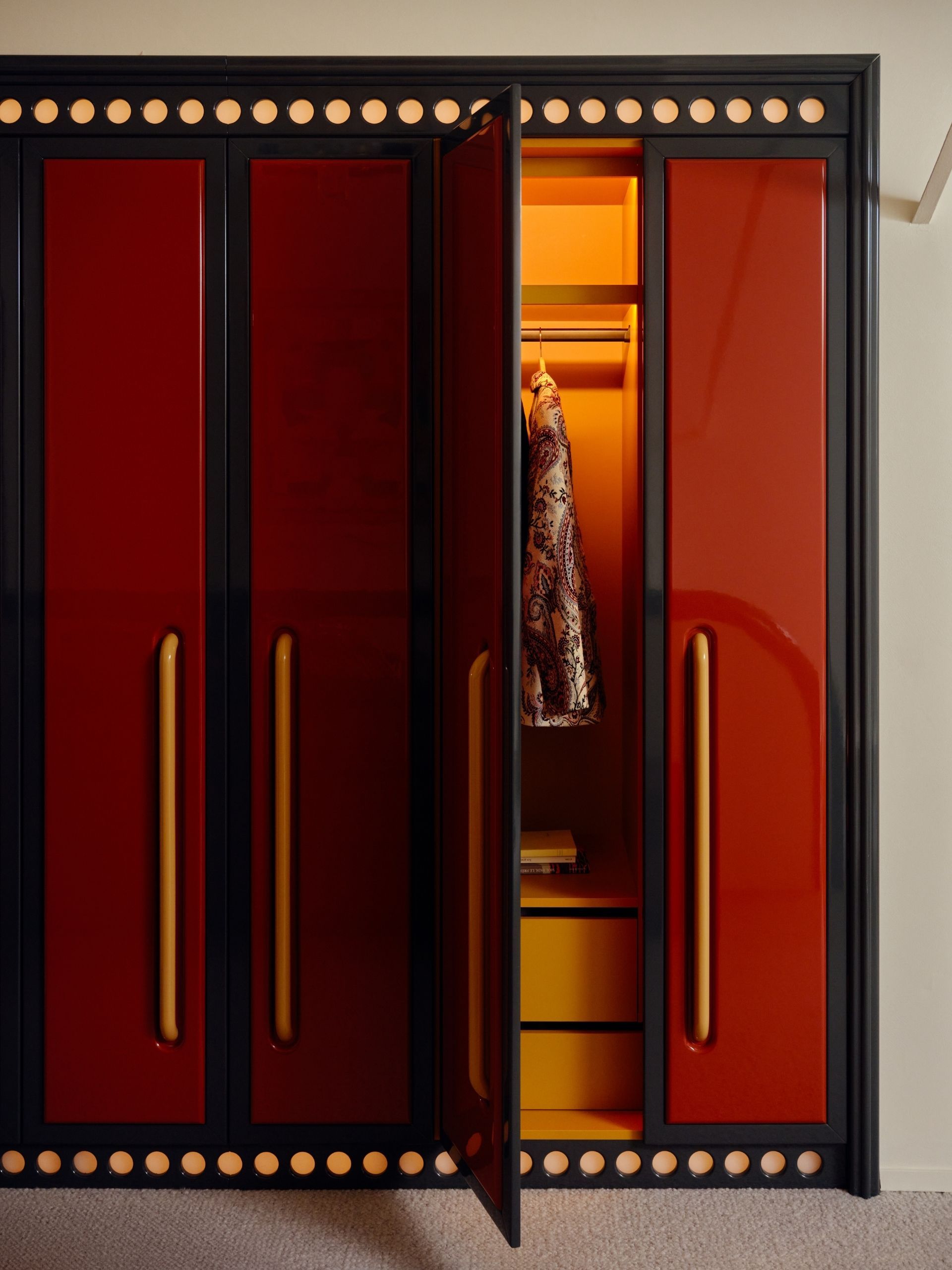 A red lacquer built-in wardrobe with yellow hardware and black trim. The door is open revealing the orange inside with lights installed.