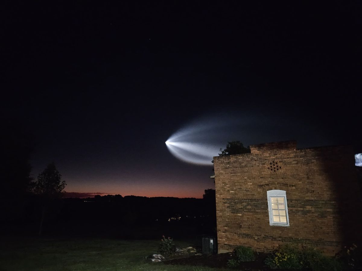 a white streak illuminated in the sky flies over a dark landscape and a brick house.