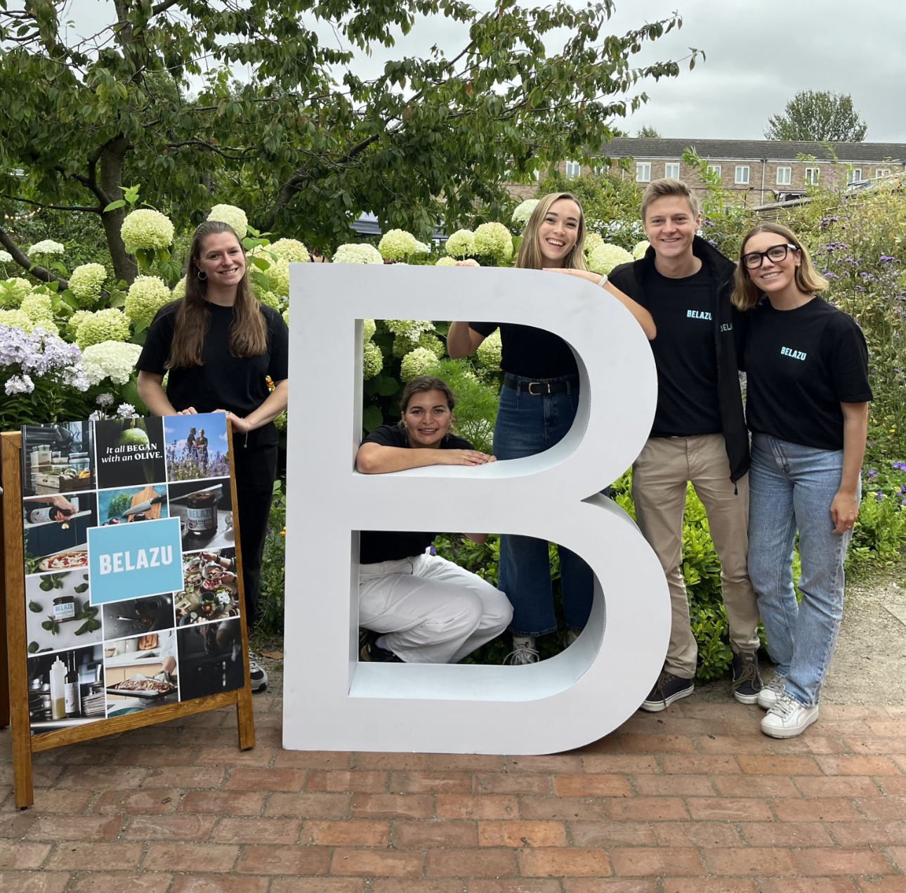 Four people posing with a large letter B, and a Belazu sign.