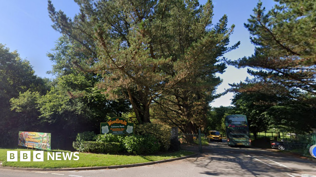 Entrance gate to Woodlands Family Theme Park. Large green trees are located either site of the site's entrance. A green Woodlands leisure park sign is located to the left of the gate. A bus is driving along the entrance road.