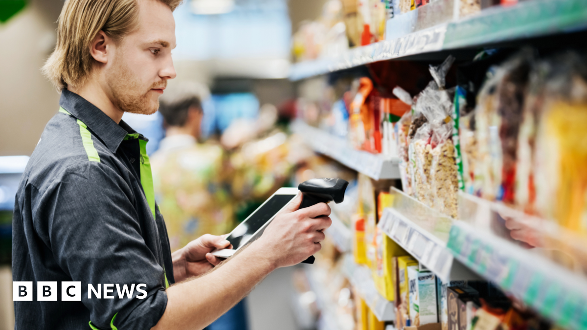 Young male supermarket worker, dressed in a dark shirt with green trimming, scans food on a shelf