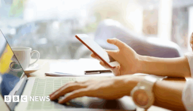 A woman uses a laptop while holding a mobile phone in her right hand.
