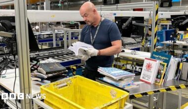 A worker in an Amazon print factory, looking at a book. There are lots of books around him. including a big yellow box. Machinery is behind him too.