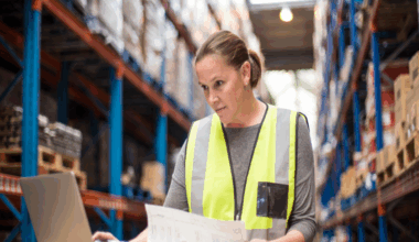 Young woman wearing yellow hi-viz vest and grey top looks at laptop while working in a warehouse