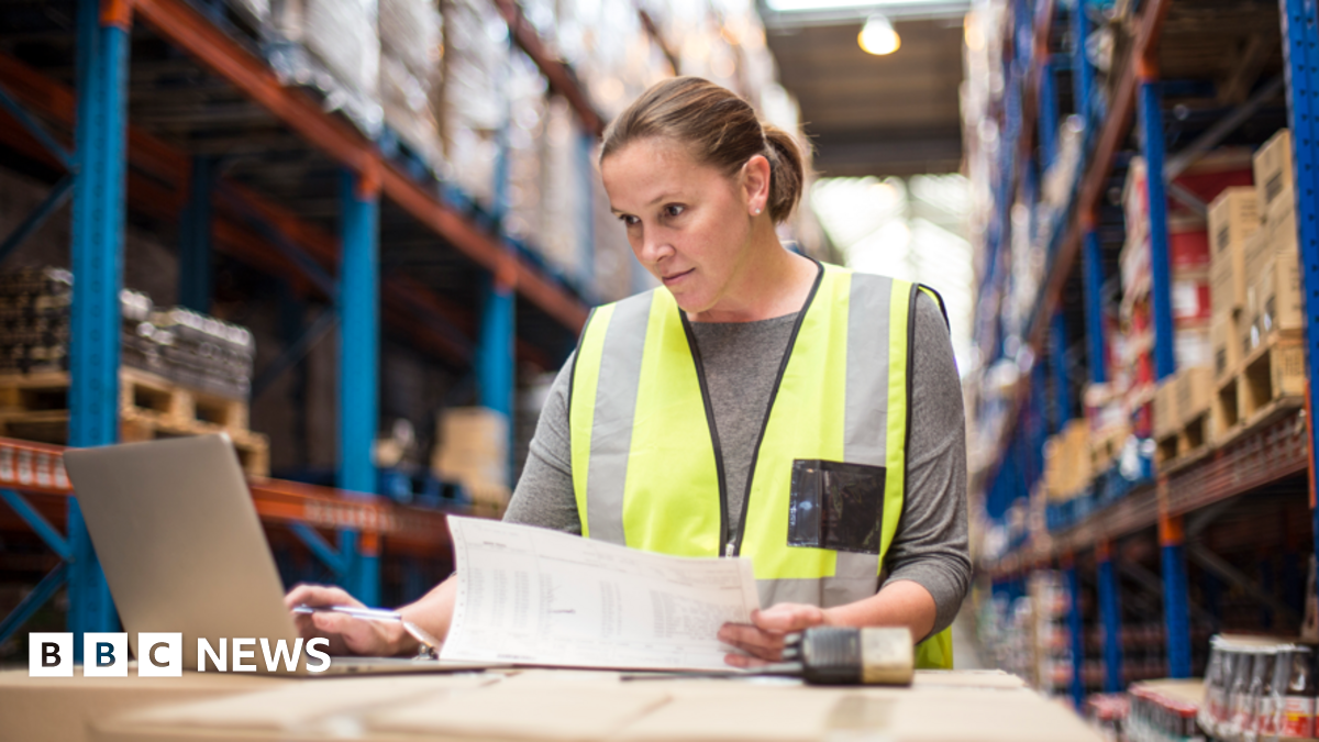 Young woman wearing yellow hi-viz vest and grey top looks at laptop while working in a warehouse